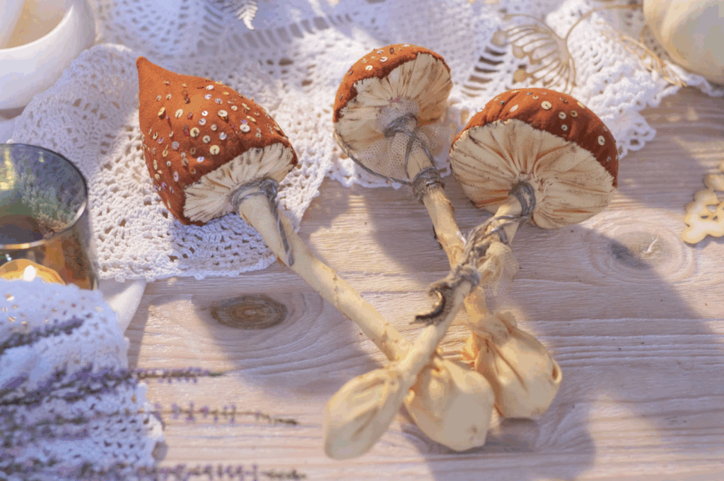 Three decorative, handcrafted fabric mushrooms with brown, sequined caps and cream-colored silk stems laid on a table with lace.